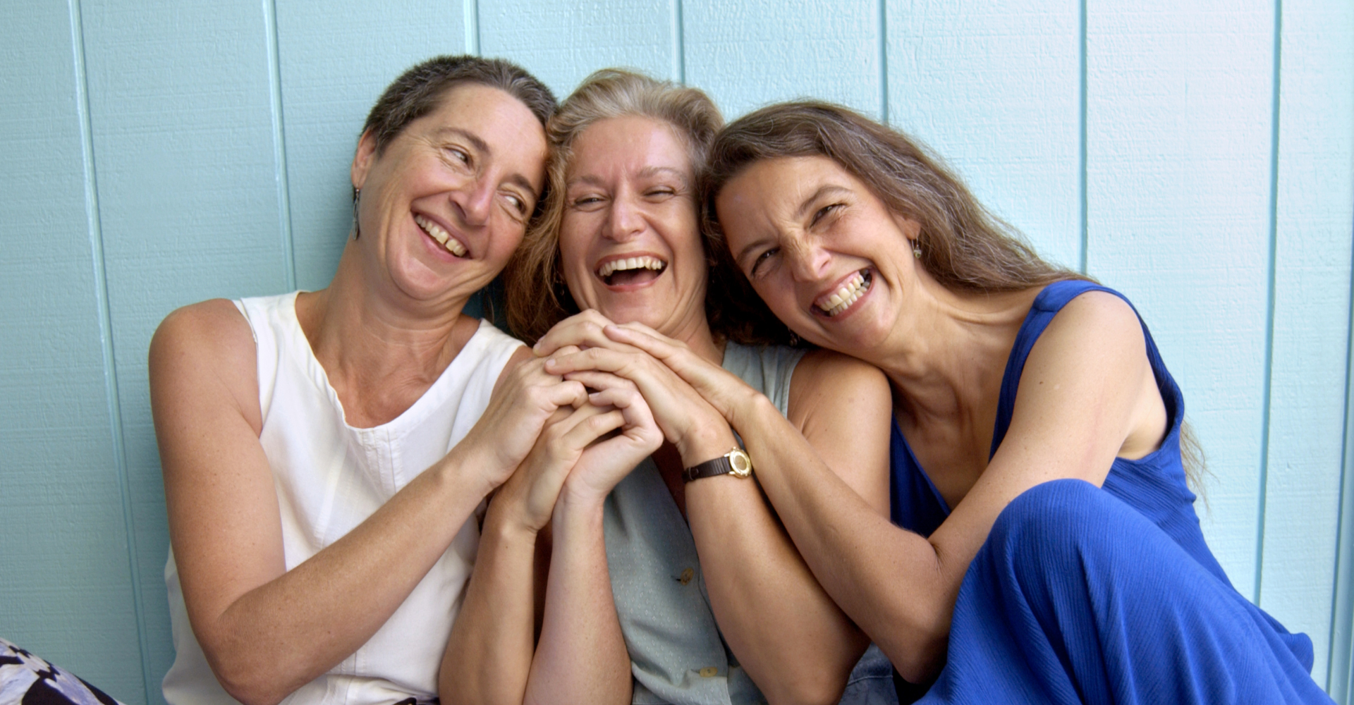 three middle aged white women who appear to be sisters, sit against a blue wall, laughing