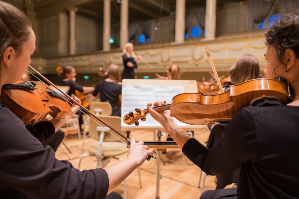 photo of string section of orchestra in rehearsal, from the back looking toward the conductor, close focus on two nearest violinists, sharing a music stand