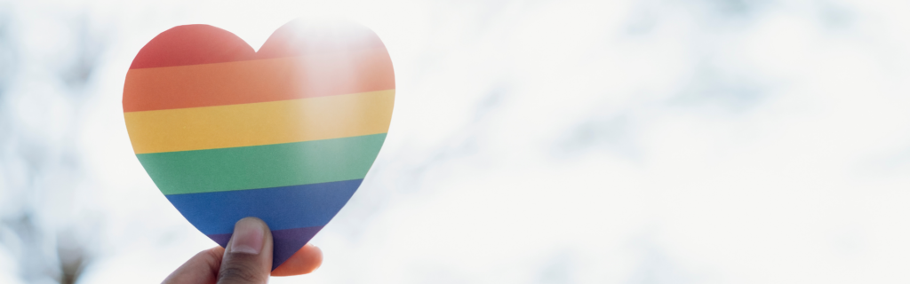 hand holding up a rainbow heart with blue sky in the background